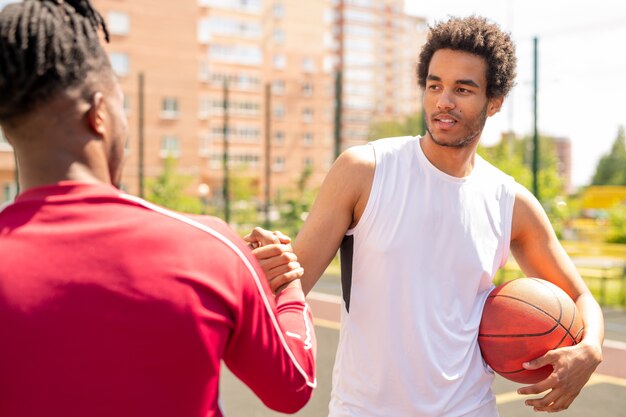 Junger interkultureller Sportler mit Ballschütteln der Hand seines Spielkameraden vor oder nach dem Basketballspiel auf dem Platz