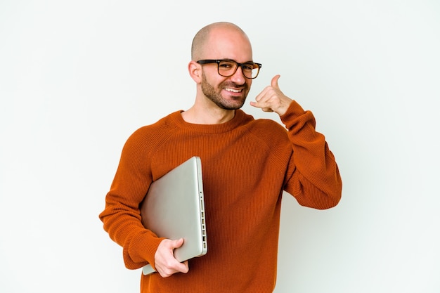 Junger kahler Mann, der einen Laptop lokalisiert auf weißer Wand hält, die eine Handy-Anrufgeste mit Fingern zeigt