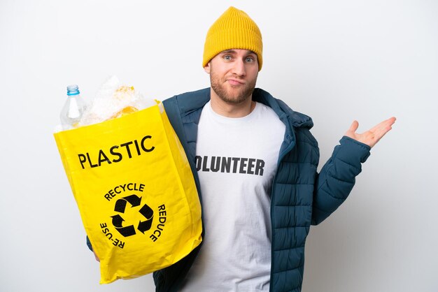 Junger Kaukasier, der eine Tasche voller Plastikflaschen hält, um sie isoliert auf weißem Hintergrund zu recyceln, und Zweifel hat, während er die Hände hebt