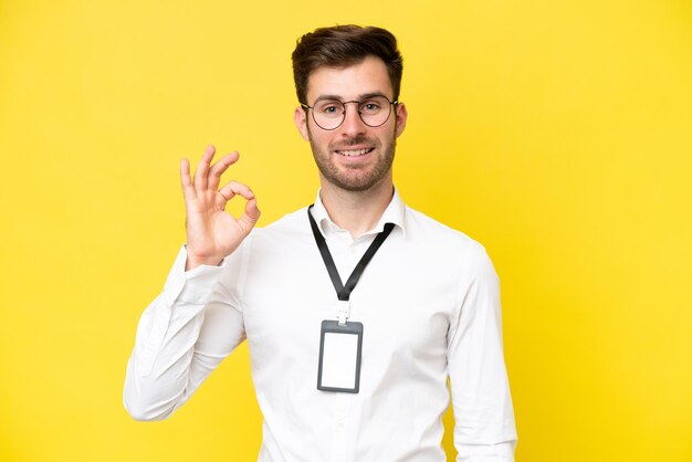 Junger Kaukasier mit ID-Karte isoliert auf gelbem Hintergrund, der mit den Fingern ein Ok-Zeichen zeigt