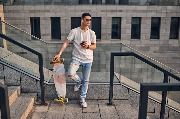 Junger kaukasischer Amerikaner mit Brille auf den Stufen mit einem Longboard vor dem Hintergrund