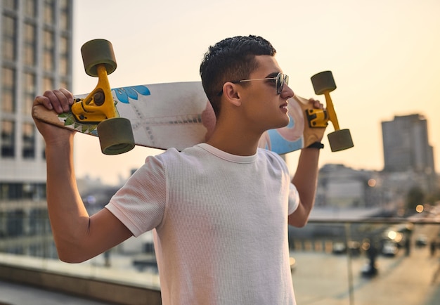 Junger kaukasischer amerikanischer Skater, der Longboard in der Hand vor dem Hintergrund der städtischen Gebäude hält