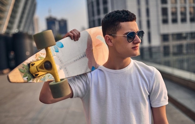 Junger kaukasischer amerikanischer Skater, der Longboard in der Hand vor dem Hintergrund der städtischen Gebäude hält
