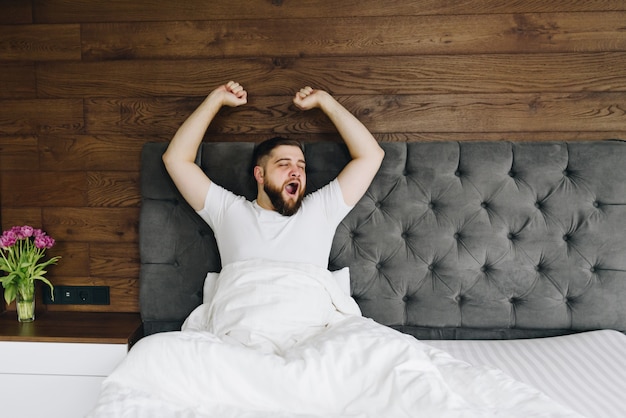 Junger kaukasischer bärtiger Mann gähnt und streckt sich in seinem hellen modernen Schlafzimmer am Morgen