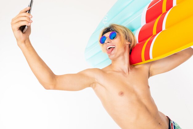 Junger kaukasischer blonder Mann steht im Badeanzug mit großer Gummi-Strandmatratze tun Selfie lokalisiert auf weißer Wand