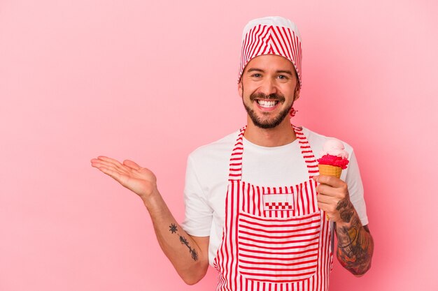 Junger kaukasischer Eismacher-Mann mit Tätowierungen, die Eiscreme einzeln auf rosafarbenem Hintergrund halten, die einen Kopienraum auf einer Handfläche zeigen und eine andere Hand an der Taille halten.