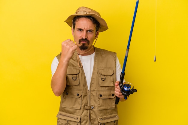 Foto junger kaukasischer fischer, der die stange isoliert auf gelbem hintergrund hält und die faust zur kamera zeigt, aggressiver gesichtsausdruck.