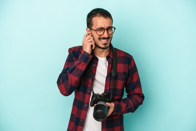 Junger kaukasischer Fotograf Mann auf blauem Hintergrund isoliert, der die Ohren mit den Händen bedeckt.