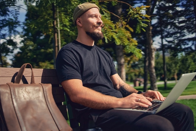 Junger kaukasischer Freiberufler sitzt mit Laptop auf der Bank im Park und genießt entspannte Freizeit