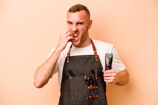 Foto junger kaukasischer friseurmann lokalisiert auf beigem hintergrund, der fingernägel beißt, nervös und sehr ängstlich