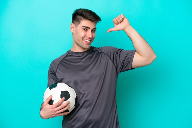 Junger kaukasischer Fußballspieler isoliert auf blauem Hintergrund stolz und selbstzufrieden