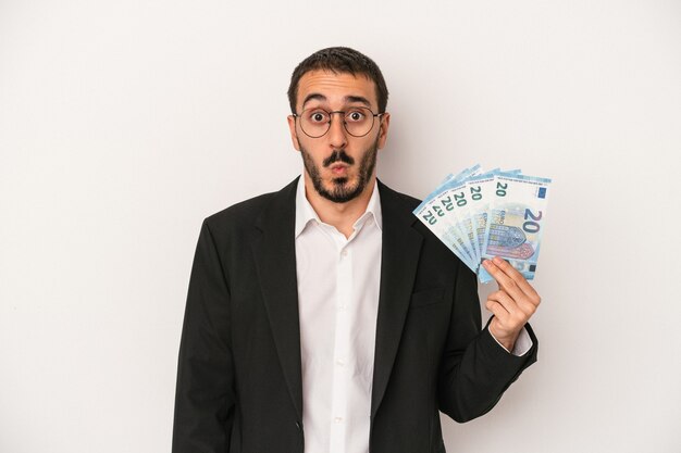Junger kaukasischer Geschäftsmann, der Banknoten lokalisiert auf weißem Hintergrund hält, zuckt mit den Schultern und öffnet die Augen verwirrt.