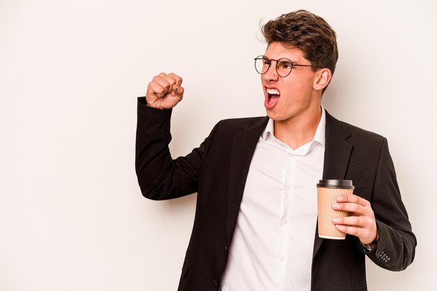 Junger kaukasischer Geschäftsmann, der Kaffee zum Mitnehmen isoliert auf weißem Hintergrund hält und nach einem Siegerkonzept die Faust hebt