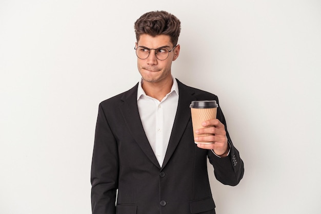 Junger kaukasischer Geschäftsmann, der Kaffee zum Mitnehmen isoliert auf weißem Hintergrund hält, verwirrt, fühlt sich zweifelnd und unsicher.