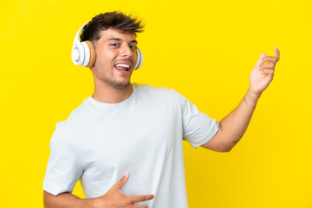 Junger kaukasischer gutaussehender Mann isoliert auf gelbem Hintergrund, der Musik hört und Gitarrengeste macht