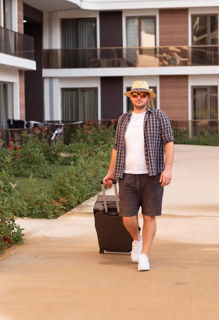 Junger kaukasischer gutaussehender Mann mit Sonnenhut-Sonnenbrille und Koffer, der zum Hotel kommtReisender Tourist im Urlaub Anfang oder Ende des Ferienkonzepts