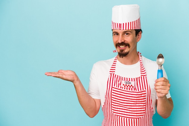 Junger kaukasischer Hersteller, der eine Schaufel isoliert auf blauem Hintergrund hält, die einen Kopienraum auf einer Handfläche zeigt und eine andere Hand an der Taille hält.