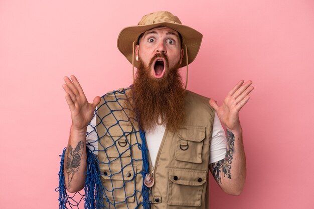 Junger kaukasischer Ingwerfischer mit langem Bart, der ein auf rosafarbenem Hintergrund isoliertes Netz hält