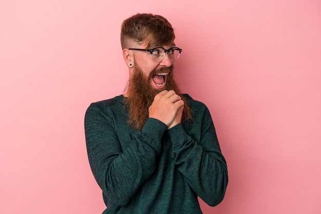 Junger kaukasischer Ingwermann mit langem Bart einzeln auf rosafarbenem Hintergrund, der um Glück betet, erstaunt und den Mund öffnet, der nach vorne schaut.