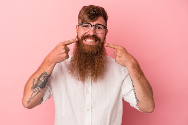 Junger kaukasischer Ingwermann mit langem Bart einzeln auf rosafarbenem Hintergrund lächelt und zeigt mit den Fingern auf den Mund.