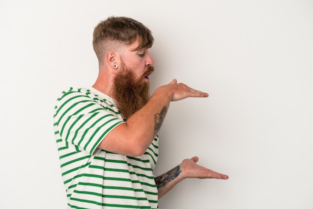 Foto junger kaukasischer ingwermann mit langem bart lokalisiert auf weißem hintergrund schockiert und überrascht, einen kopienraum zwischen den händen zu halten.