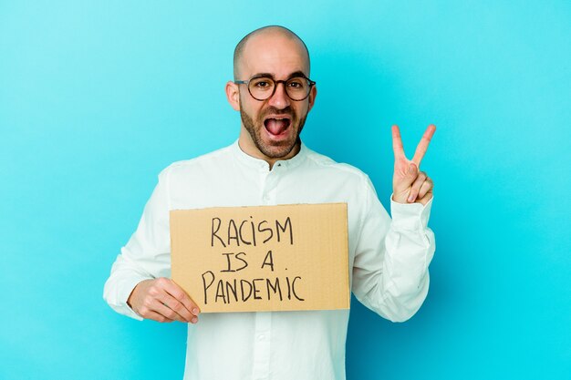 Junger kaukasischer kahlköpfiger Mann, der einen Rassismus hält, ist eine Pandemie, die auf der weißen Wand freudig und sorglos isoliert ein Friedenssymbol mit den Fingern zeigt