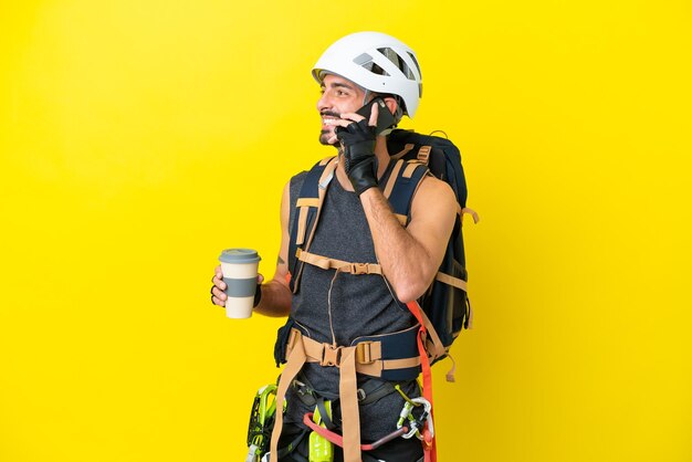 Junger kaukasischer Kletterer isoliert auf gelbem Hintergrund mit Kaffee zum Mitnehmen und einem Handy