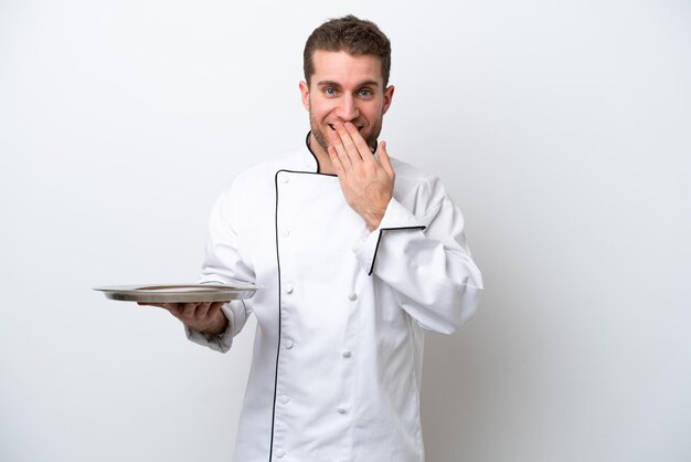 Junger kaukasischer Koch mit Tablett isoliert auf weißem Hintergrund glücklich und lächelnd, den Mund mit der Hand bedeckend