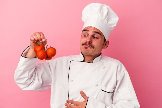 Junger kaukasischer Kochmann, der Tomaten lokalisiert auf rosa Hintergrund hält