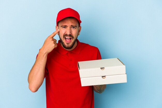 Junger kaukasischer Lieferbote mit Tätowierungen, die Pizzas einzeln auf blauem Hintergrund halten, die eine Enttäuschungsgeste mit dem Zeigefinger zeigen.