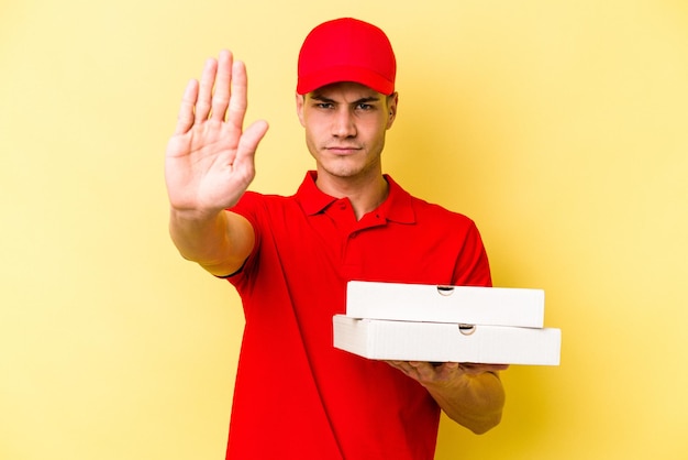 Junger kaukasischer Liefermann, der Pizzen isoliert auf gelbem Hintergrund hält und mit ausgestreckter Hand mit Stoppschild steht und Sie daran hindert.
