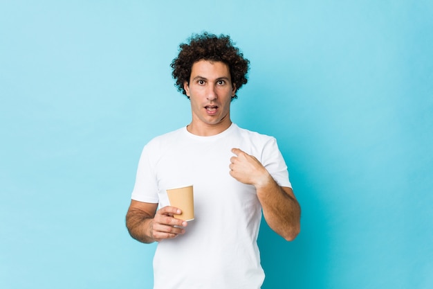 Junger kaukasischer lockiger Mann, der einen Kaffee zum Mitnehmen hält, zeigte überrascht auf sich selbst und lächelte breit.