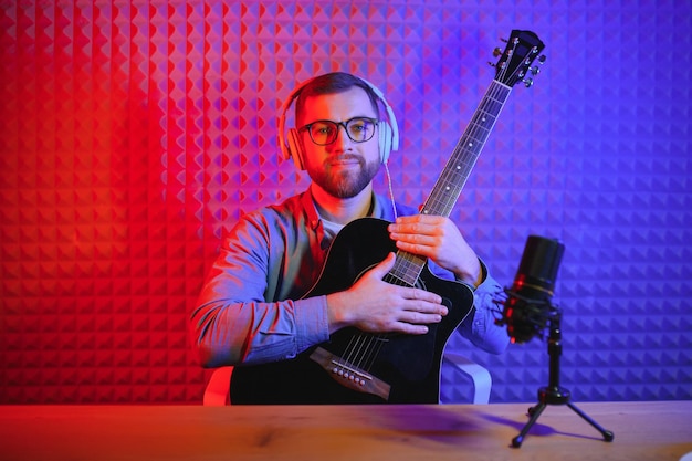 Junger kaukasischer männlicher Sänger in Kopfhörern hält Gitarre spielen Rekord neue Single auf dem Smartphone im Heimstudio Millennial Man Künstler verwenden Musikinstrumente singen Musikvideos auf Handykamera aufnehmen