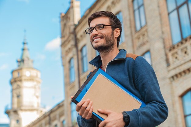 Junger kaukasischer männlicher Student mit Büchern draußen in der Nähe des Campus