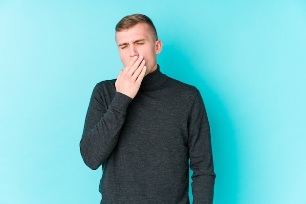Junger kaukasischer Mann auf einer blauen Wand, die gähnt und eine müde Geste zeigt, die Mund mit Hand bedeckt.