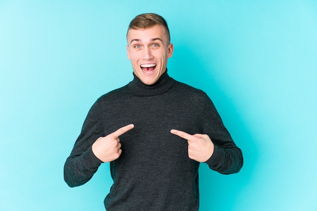 Junger kaukasischer Mann auf einer blauen Wand überrascht, mit dem Finger zeigend, breit lächelnd.