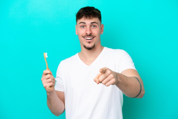 Junger kaukasischer Mann beim Zähneputzen isoliert auf blauem Hintergrund überrascht und nach vorne zeigend