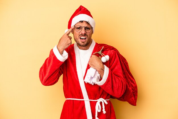 Junger kaukasischer Mann, der als Weihnachtsmann verkleidet ist, isoliert auf gelbem Hintergrund, der eine Enttäuschungsgeste mit dem Zeigefinger zeigt.