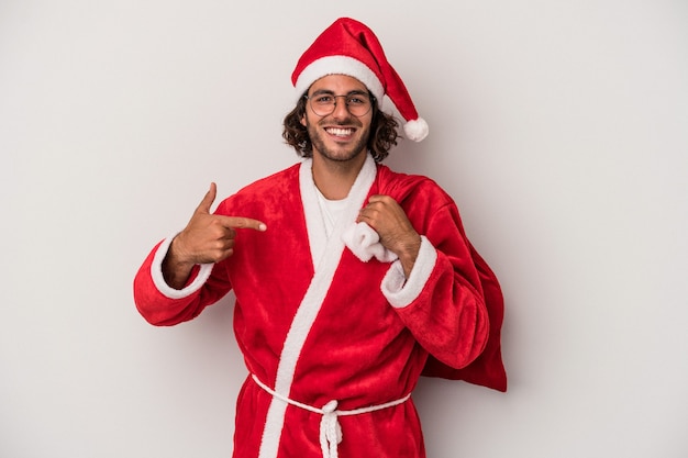 Junger kaukasischer Mann, der als Weihnachtsmann verkleidet ist, isoliert auf grauem Hintergrund, der mit der Hand auf einen Hemdkopierraum zeigt, stolz und selbstbewusst