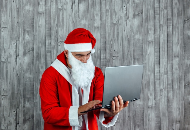 Junger kaukasischer Mann, der als Weihnachtsmann verkleidet ist und eine Internetsuche macht. Konzept des Online-Shoppings auf dem Laptop