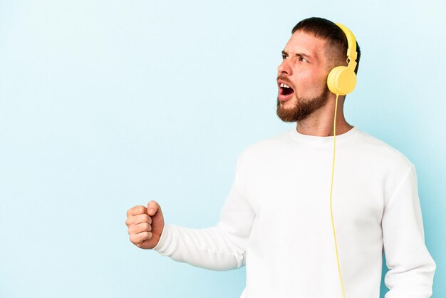 Junger kaukasischer Mann, der auf blauem Hintergrund Musik hört, die nach einem Sieg die Faust anhebt, Gewinnerkonzept.