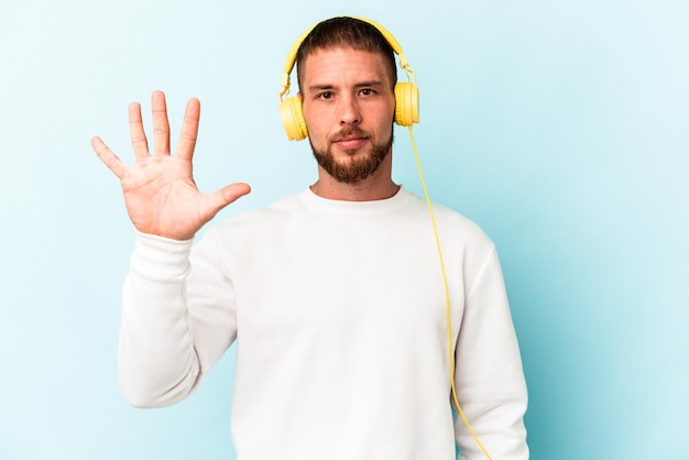 Junger kaukasischer Mann, der auf blauem Hintergrund Musik hört, lächelt fröhlich und zeigt Nummer fünf mit den Fingern.