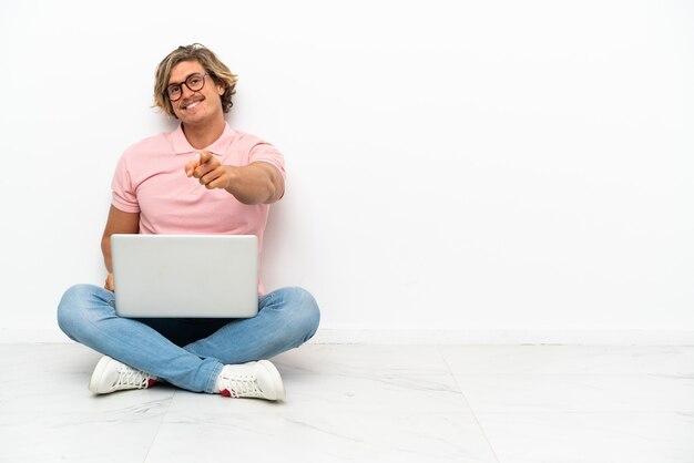 Junger kaukasischer Mann, der auf dem Boden mit seinem Laptop lokalisiert auf weißem Hintergrund sitzt, der vorne mit glücklichem Ausdruck zeigt