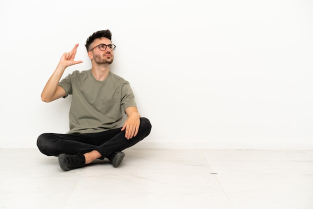 Junger kaukasischer Mann, der auf dem Boden sitzt, isoliert auf weißem Hintergrund, mit gekreuzten Fingern und das Beste wünscht
