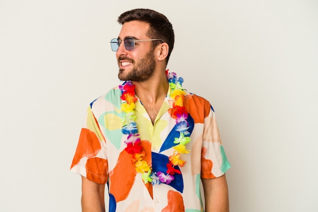 Junger kaukasischer Mann, der auf einer hawaiianischen Party tanzt, isoliert auf weißem Hintergrund, sieht beiseite lächelnd, fröhlich und angenehm aus.