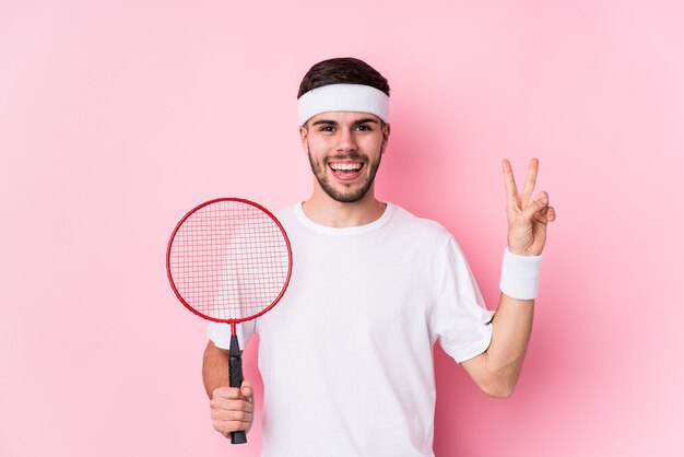 Junger kaukasischer Mann, der Badminton spielt, isolierte freudig und sorglos, ein Friedenssymbol mit den Fingern zeigend.