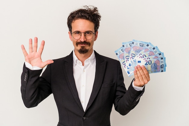 Junger kaukasischer Mann, der Banknoten hält, die auf weißem Hintergrund lokalisiert werden, lächelt fröhlich und zeigt Nummer fünf mit den Fingern.