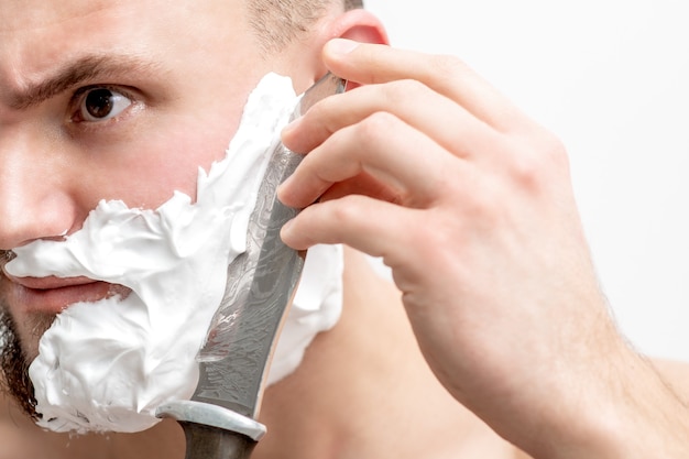 Junger kaukasischer Mann, der Bart mit Messer auf weißem Hintergrund rasiert