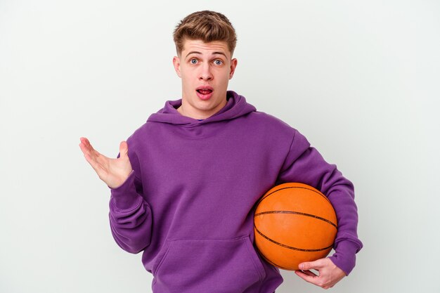 Junger kaukasischer Mann, der Basketball isolierte Wand spielt, überrascht und schockiert.