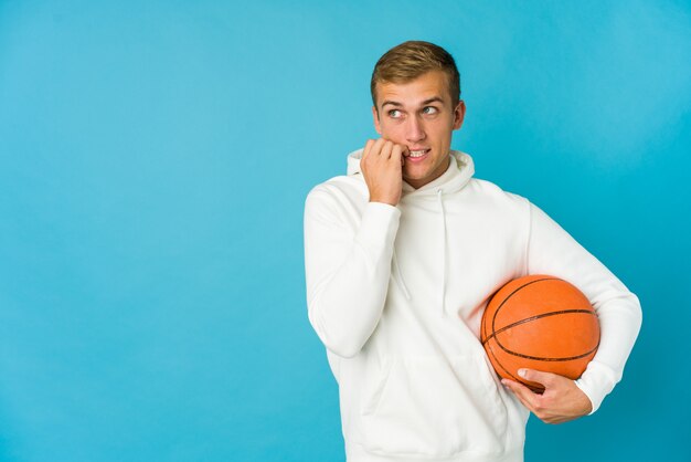 Junger kaukasischer Mann, der Basketball spielt, isoliert auf blau entspanntem Denken über etwas, das einen Kopienraum betrachtet.